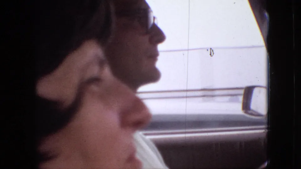 Inside a moving car in the supplied 1968 Branson, Missouri clip, a dark-haired passenger is seen in soft profile while a driver wearing glasses appears beyond her.