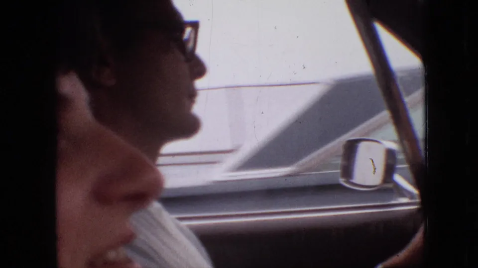 The driver, wearing glasses, appears in profile inside the car while the passenger's face fills the foreground, with the side mirror and exterior vehicle body visible through the window.