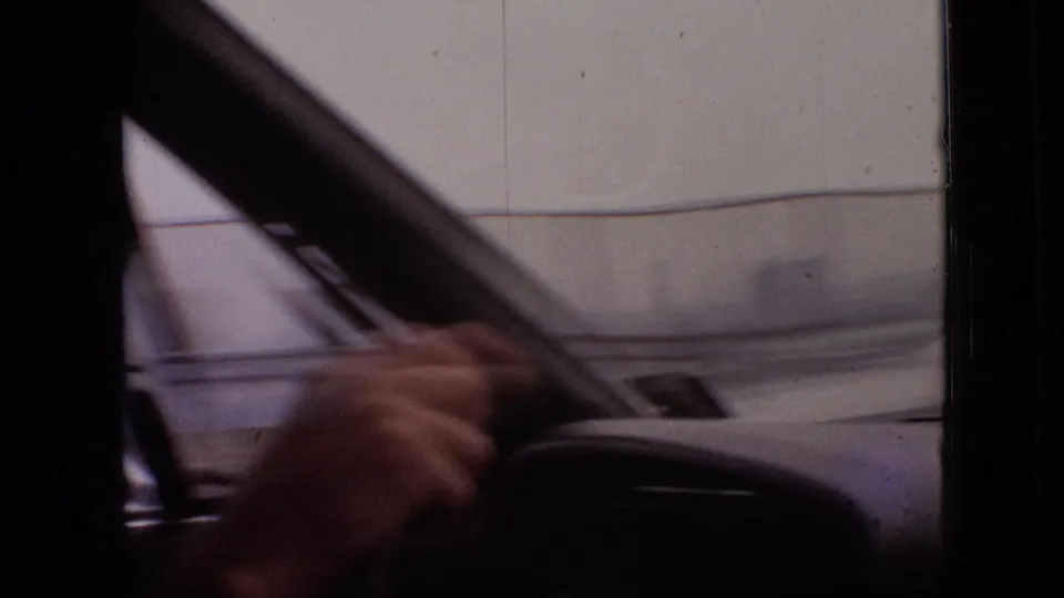 A close interior view shows the driver holding or reaching near the steering wheel, with the passenger partly visible and blurred exterior surfaces passing outside the windshield.