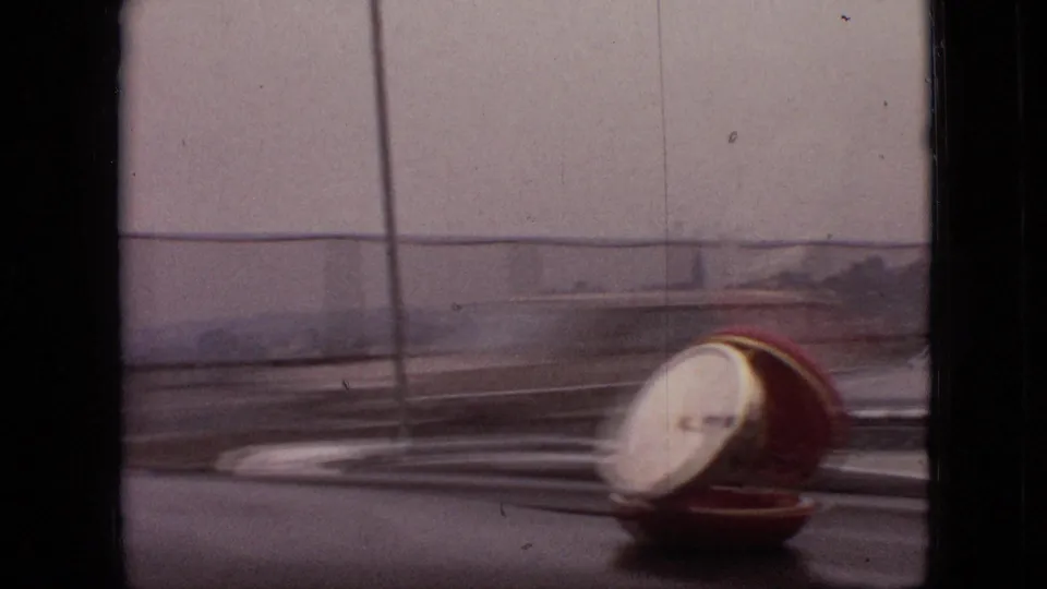 A red-and-white dashboard clock sits near the windshield inside the car, with vertical window framing and a blurred road or vehicle surface outside.