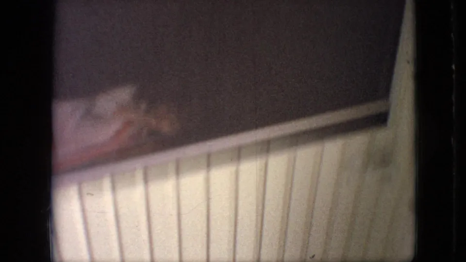 1968 Branson, Missouri home-movie frame showing pale vertical slats beneath a darker window area, with a blurred chair-like object or household item at the left edge.