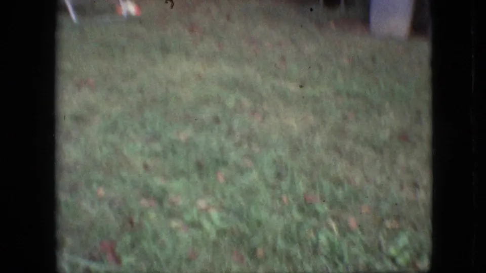 Soft 1968 Branson 8mm frame with grass in the foreground, a blurred person near the top edge, and pale containers in the background.