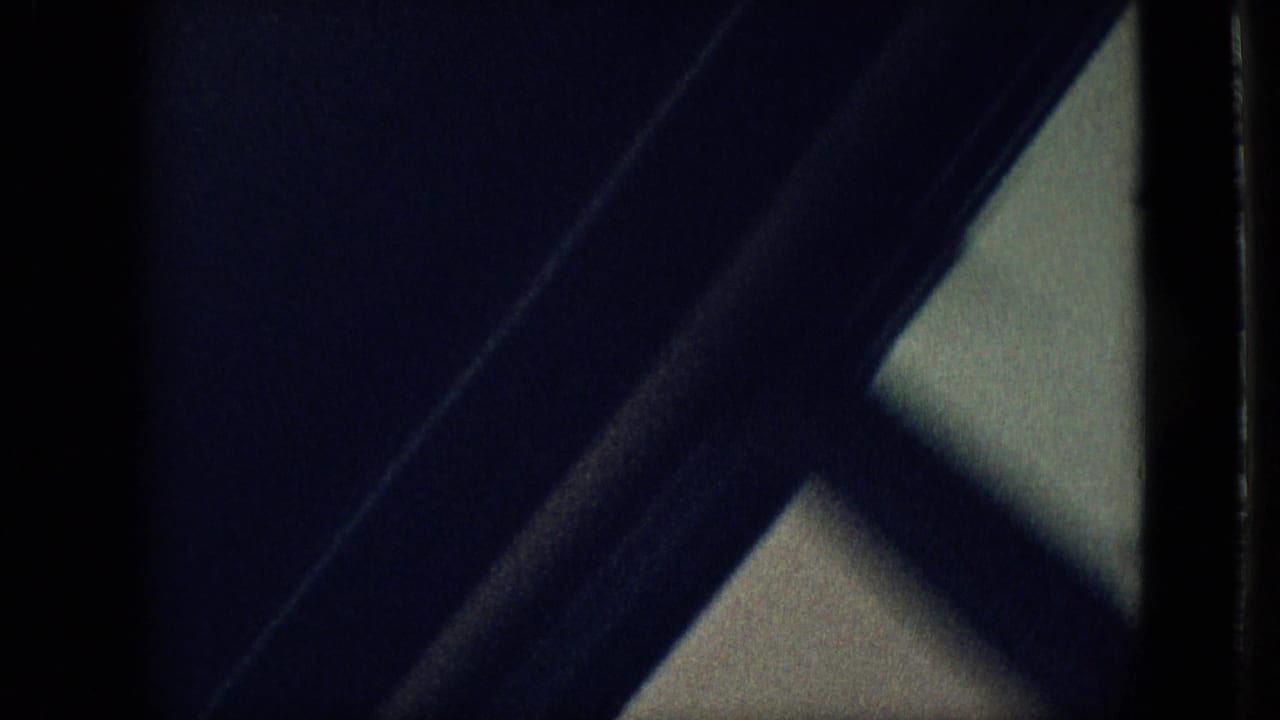 Dark underexposed 8mm frame from the Branson home movie, showing a diagonal frame or beam and a pale window-like area at the edge.