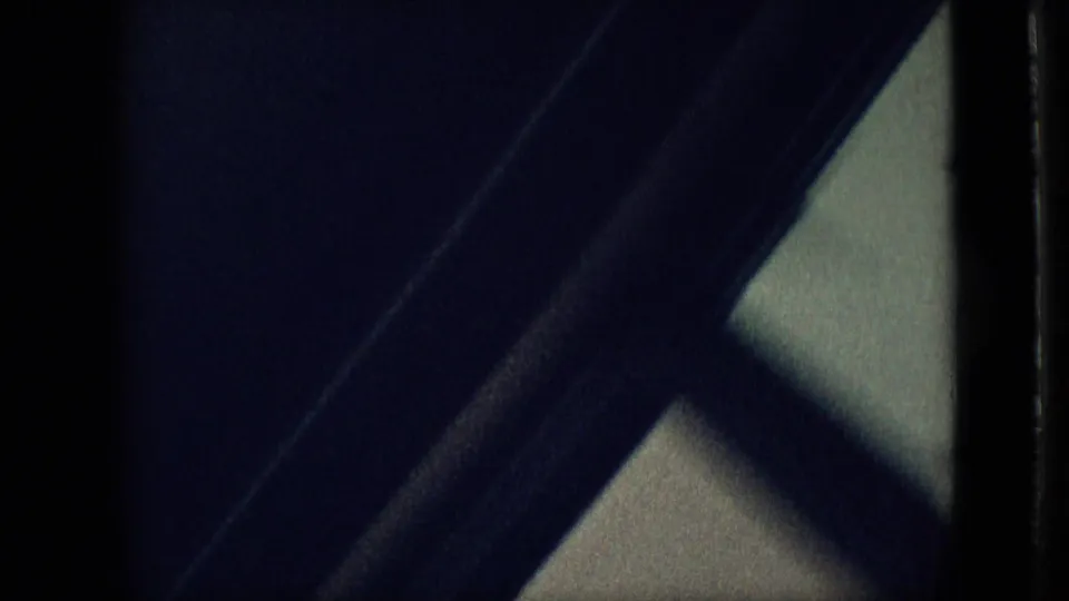 Dark underexposed 8mm frame from the Branson home movie, showing a diagonal frame or beam and a pale window-like area at the edge.