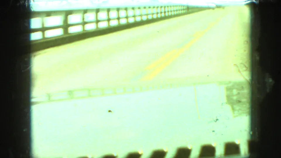 Bright 1968 8mm travel frame from Branson, Missouri, looking from inside a moving vehicle across a bridge or roadway with railings and a yellow center line.