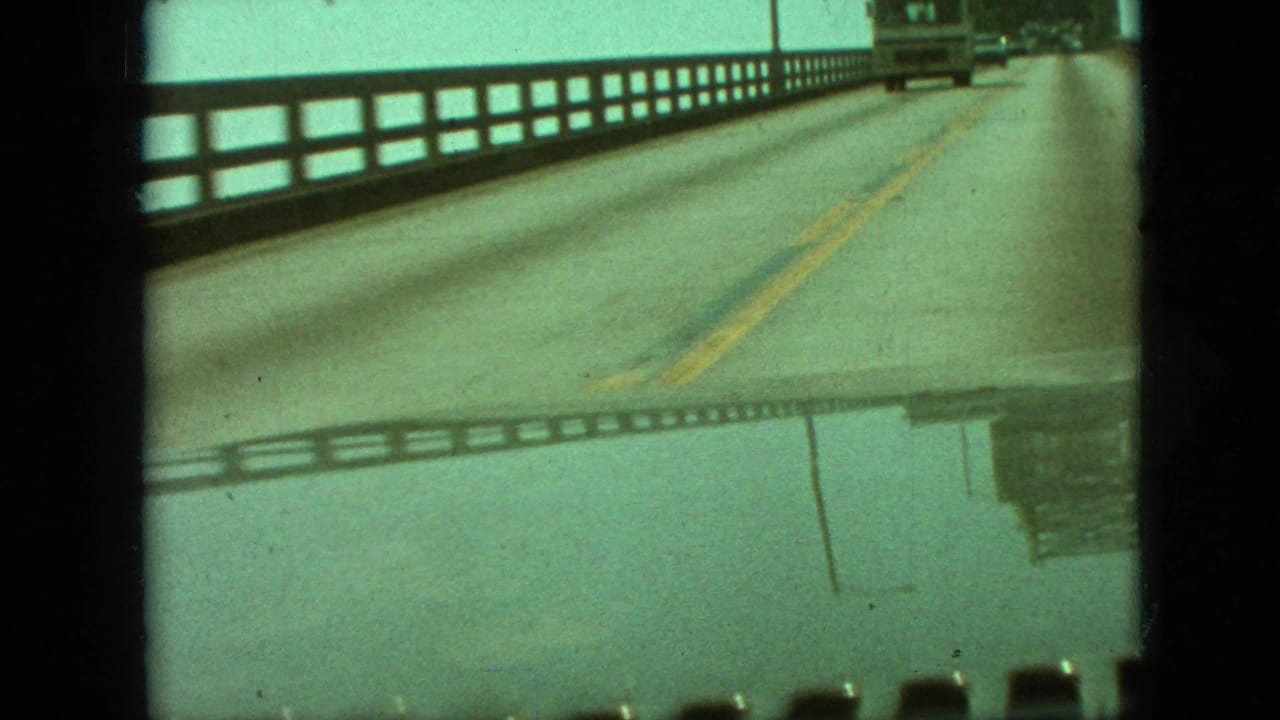 1968 Branson, Missouri home-movie frame showing a forward view from a moving vehicle on a two-lane bridge with a side railing and yellow center stripe.