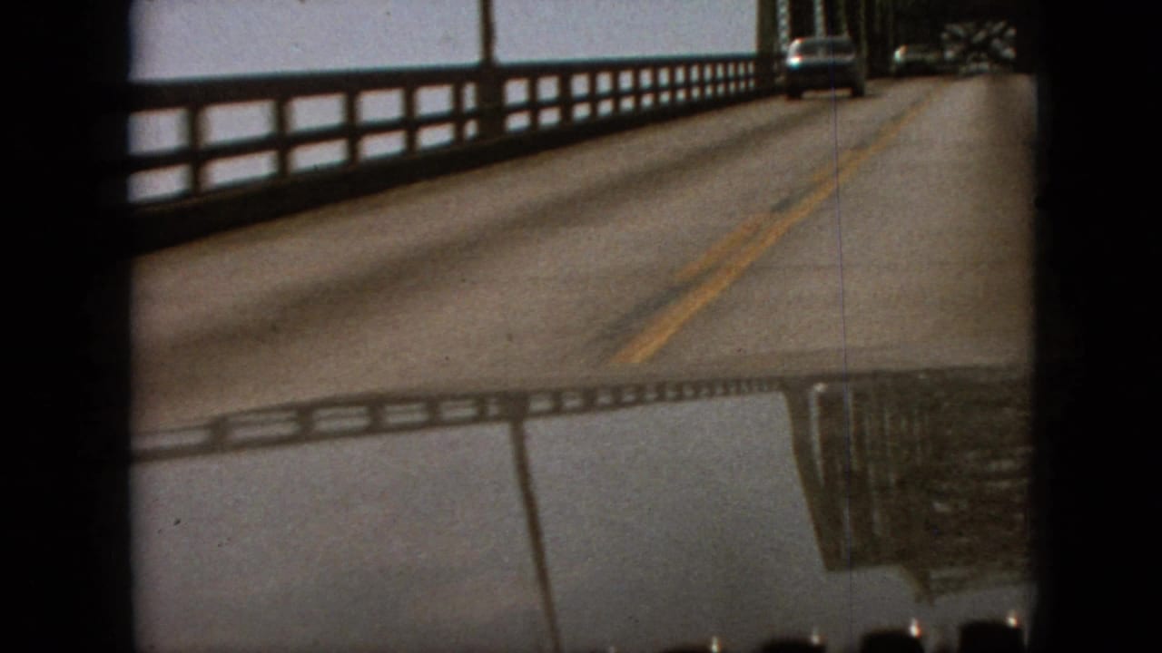 1968 Branson, Missouri home-movie frame showing a moving-car view along a wet steel truss bridge road, with yellow center lines and dark film-frame borders.