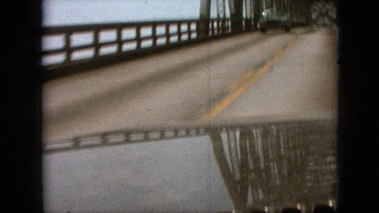 Wet bridge roadway seen from a car, with railings, center lines, and reflected bridge structure.