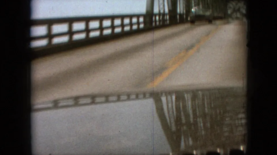 Moving-car home-movie view on a wet bridge road in Branson, Missouri, with steel railings, yellow lane markings, and reflected bridge structure in the lower frame.