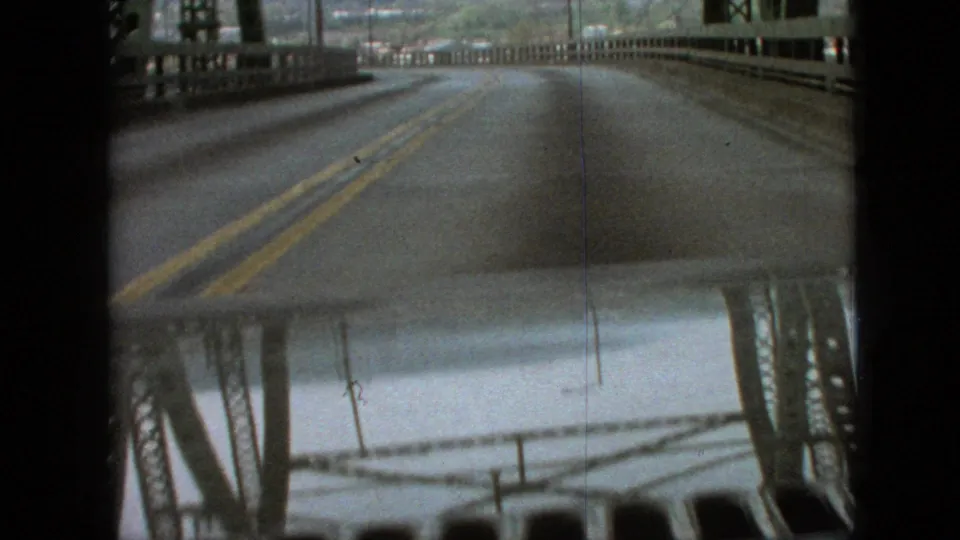 Moving-car view across a wet two-lane bridge road in 1968 Branson, Missouri, with yellow center lines, steel bridge sides, and reflections near the bottom of frame.