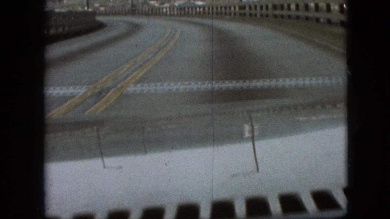 The moving car crosses a bridge on a curving two-lane road, with muted 8mm color, bridge railings, and the vehicle hood visible in the foreground.