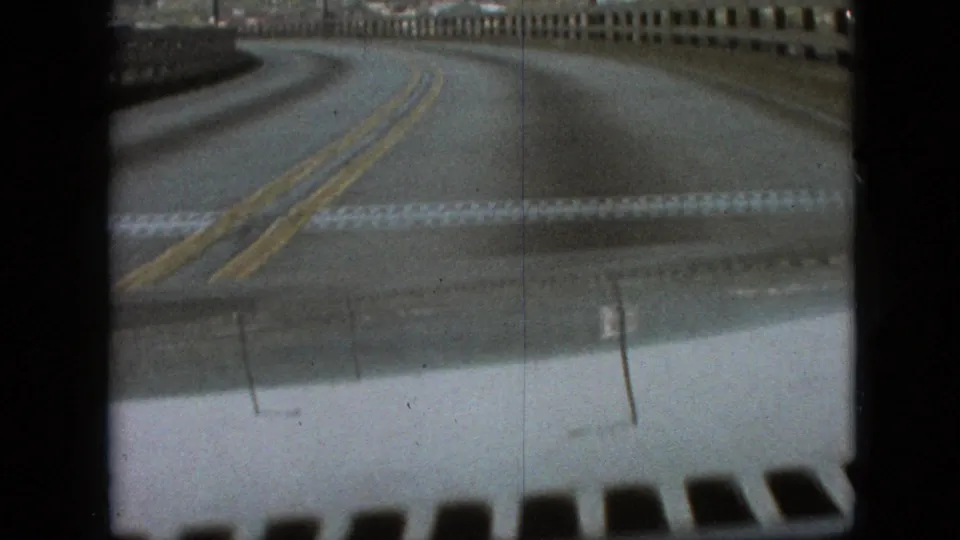 The moving car crosses a bridge on a curving two-lane road, with muted 8mm color, bridge railings, and the vehicle hood visible in the foreground.