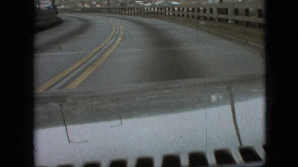 A driver POV view shows the road bending left across the bridge, with double yellow lines, railings, and small-gauge film grain visible throughout the frame.