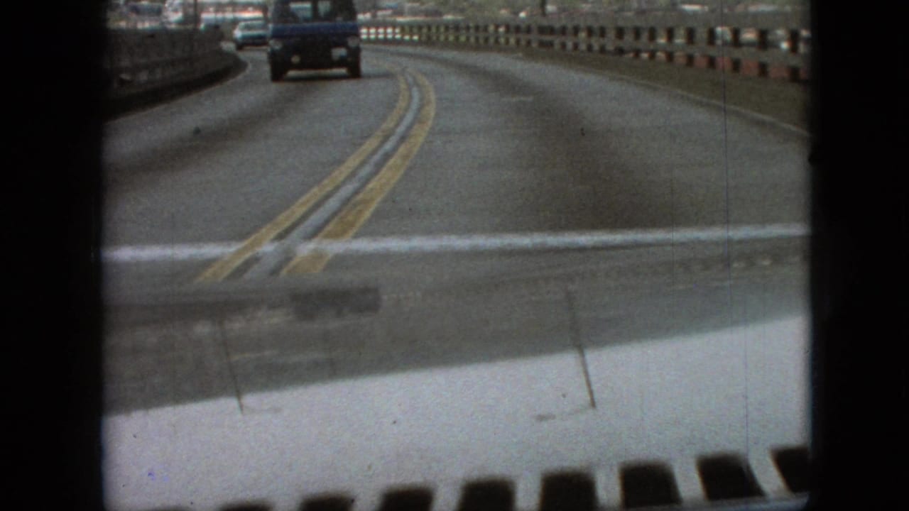 1968 Branson, Missouri 8mm frame showing a first-person car view on a bridge, with double yellow road markings, concrete guardrail, and vehicles ahead.
