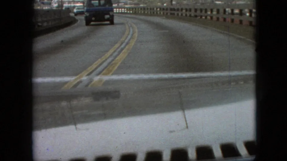 1968 Branson, Missouri 8mm frame showing a first-person car view on a bridge, with double yellow road markings, concrete guardrail, and vehicles ahead.