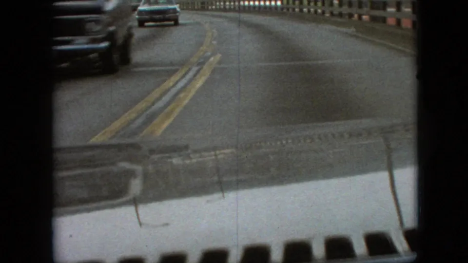 First-person 8mm travel view on a Branson, Missouri bridge in 1968, with double yellow center lines, guardrail, and nearby passing traffic.