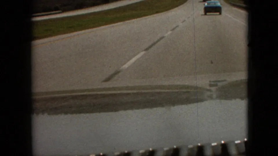 1968 Branson, Missouri home-movie frame from a moving car, showing a blue vehicle ahead on a two-lane highway with grass shoulder and dashboard edge visible.