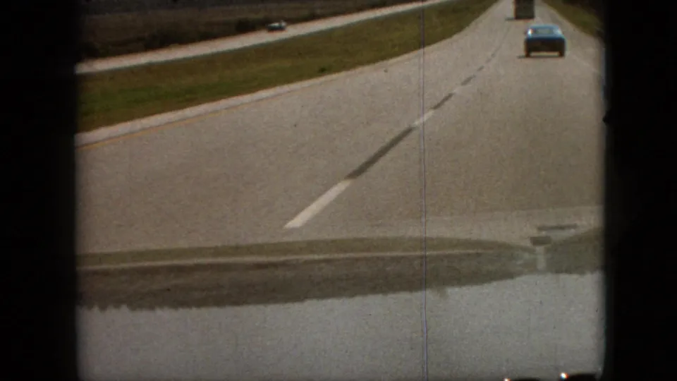 First-person 8mm road-trip frame from Branson, Missouri in 1968, looking down a highway toward a blue vehicle with open roadside grass and visible film-frame edges.