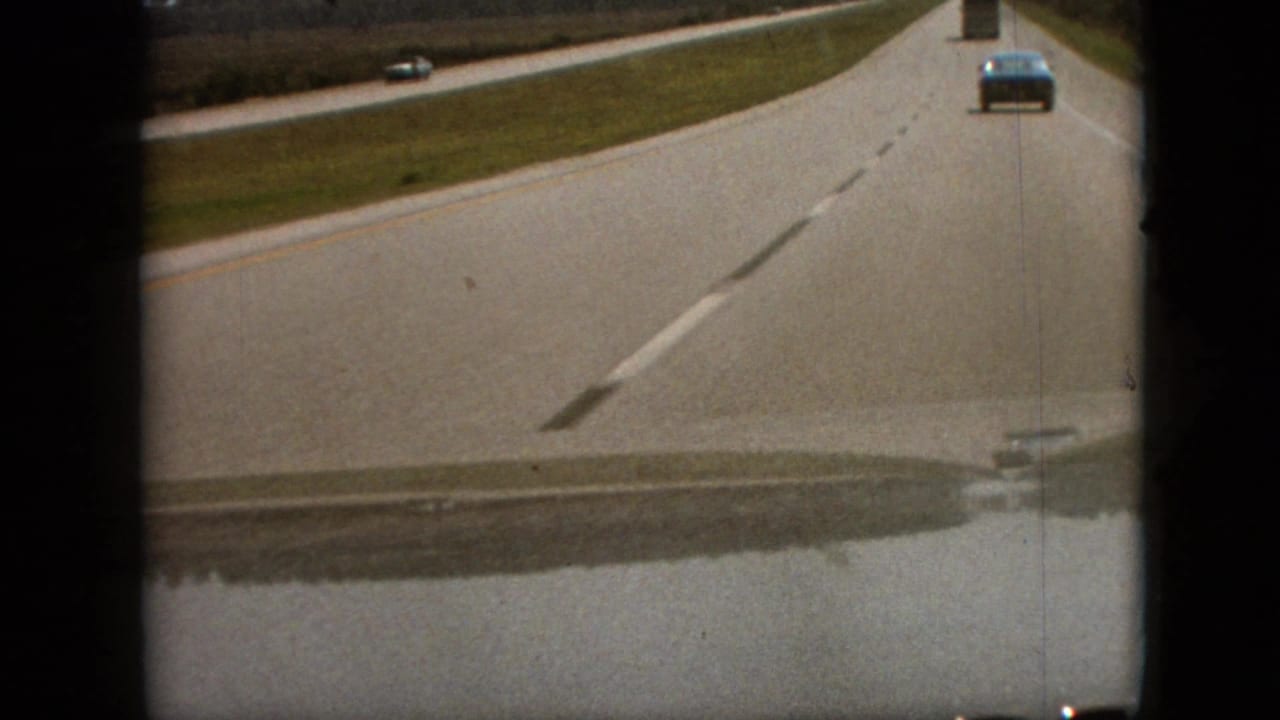 1968 8mm road-travel frame near Branson, Missouri, showing a passenger-view highway scene with a car ahead, faded lane lines, grass shoulders, and dark film gate edges.