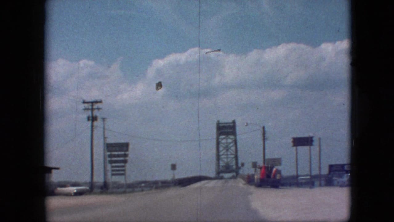 1968 8mm frame near Branson, Missouri, showing the road leading toward a steel truss bridge with roadside signs, utility poles, a vehicle, and bright clouds overhead.