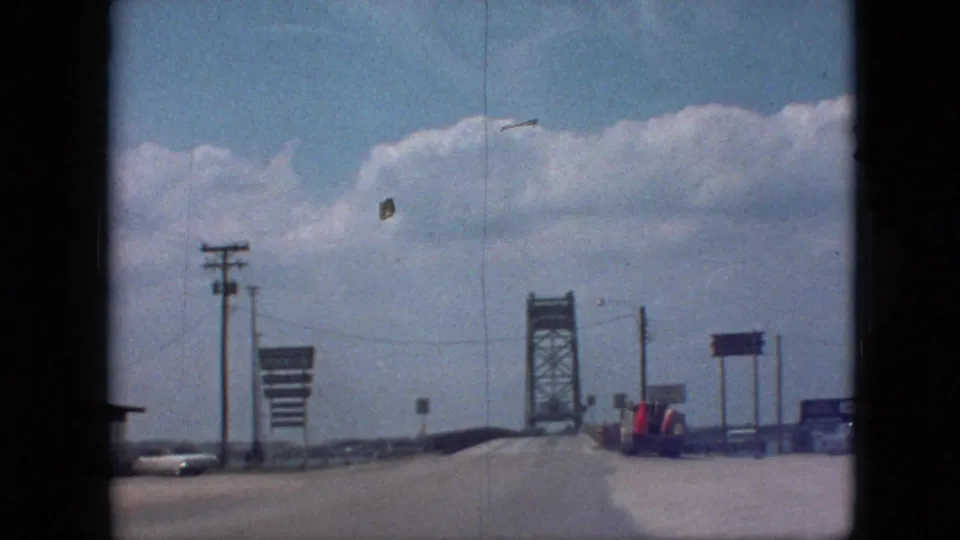 1968 8mm frame near Branson, Missouri, showing the road leading toward a steel truss bridge with roadside signs, utility poles, a vehicle, and bright clouds overhead.