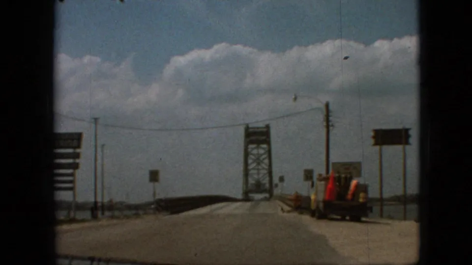 1968 8mm bridge-approach frame near Branson, Missouri, with a truck or work vehicle at the right shoulder, roadside signs, utility poles, and a steel truss bridge ahead.