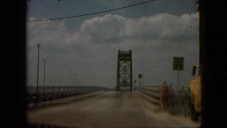 1968 8mm frame near Branson, Missouri, showing the roadway and guardrails leading toward a steel truss bridge, with water to the side, roadside signs, and a speed sign visible.