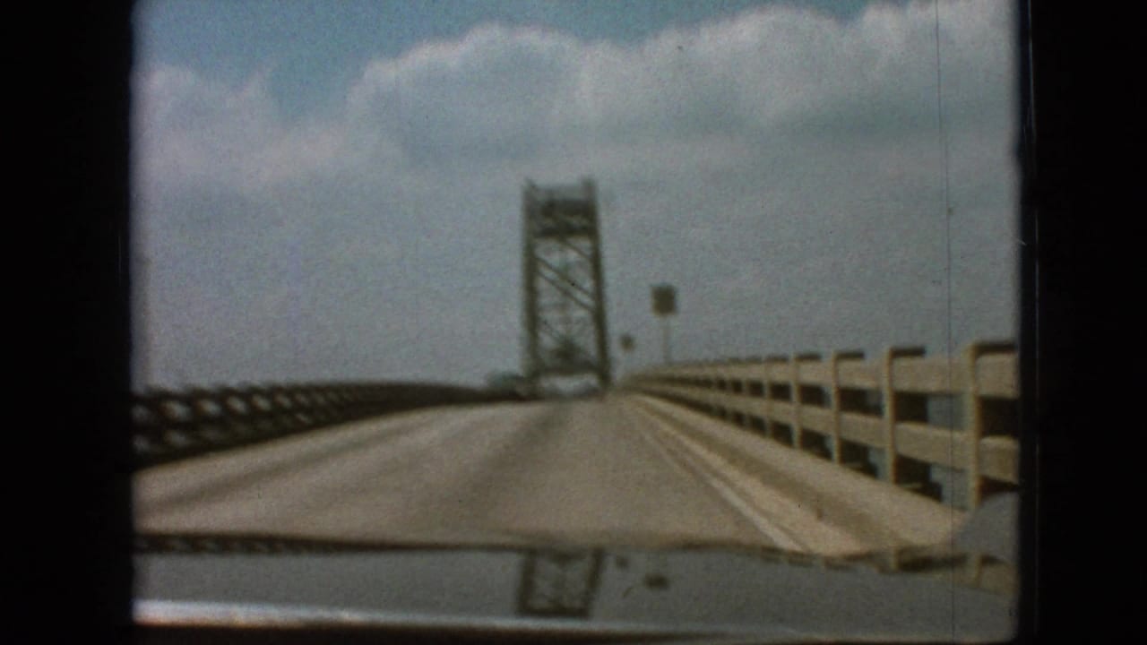 1968 home-movie view recorded as Branson, Missouri, showing a car traveling across a long bridge toward a steel truss tower under a partly cloudy sky.