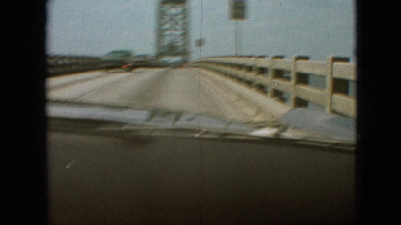 Bridge deck and guardrails seen through a car windshield near a steel tower.