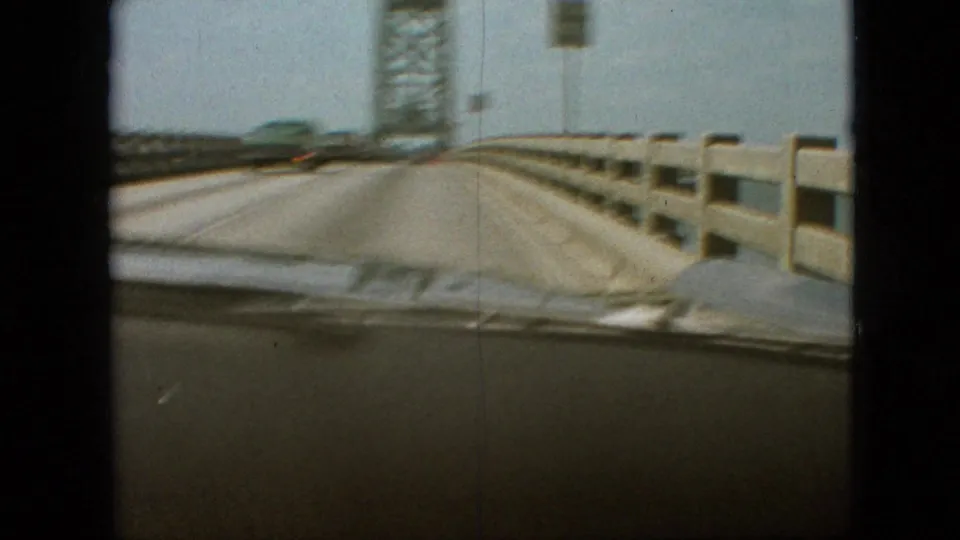 Windshield-framed 8mm view of the bridge deck, roadside railings, and steel tower as the vehicle continues across the span.