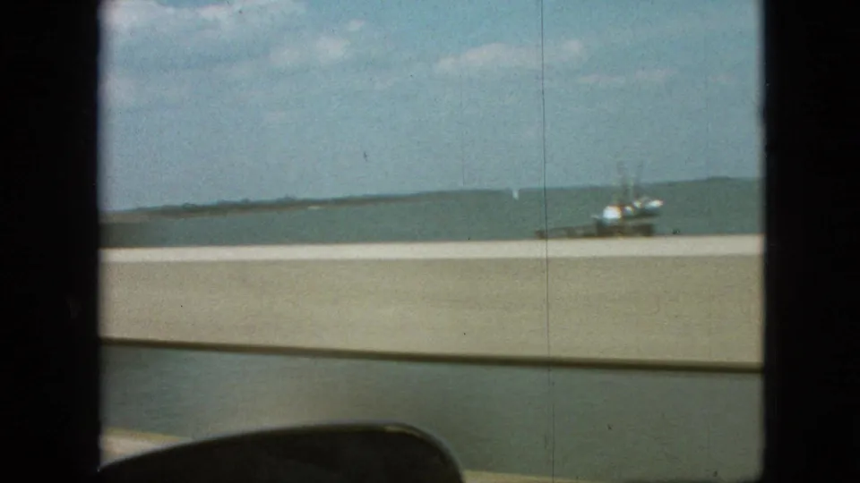 End frame shows a large body of water, low shoreline, bridge barrier, windshield edge, and a small boat-like vessel during the bridge crossing.