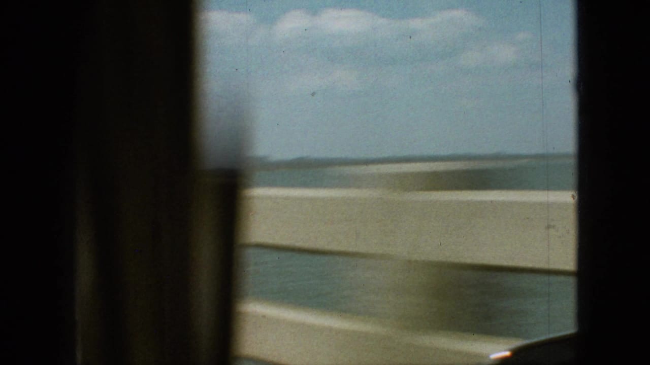 1968 home-movie frame from the Branson, Missouri clip showing a blurred car-window view of a waterside roadway, pale guard edge, distant shoreline, and cloudy sky.
