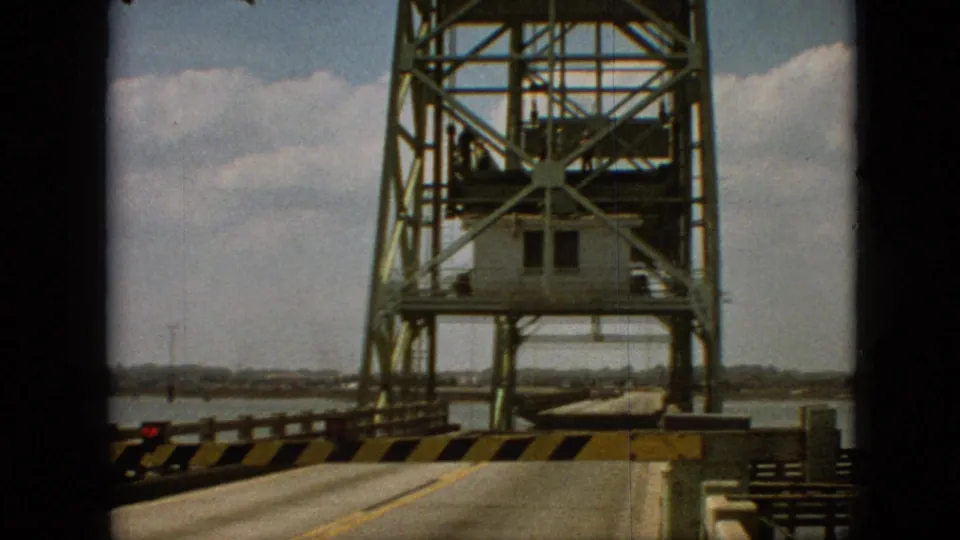 Final 1968 Branson, Missouri frame showing the stopped roadway leading toward a raised green steel bridge tower, striped barrier, control cabin, water, cloudy sky, and distant shore.
