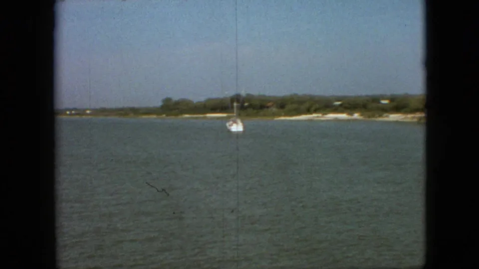 1968 Branson-area home-movie frame showing a small white sailboat on the water with a low shoreline and trees beyond it.