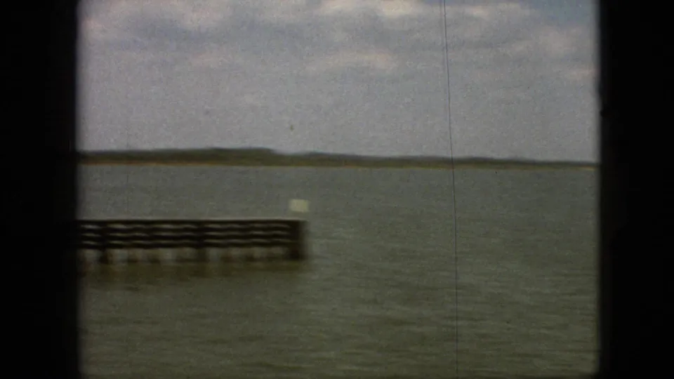 1968 Branson-area home-movie frame showing a low dock or pier over open water with a distant wooded shoreline under a cloudy sky.