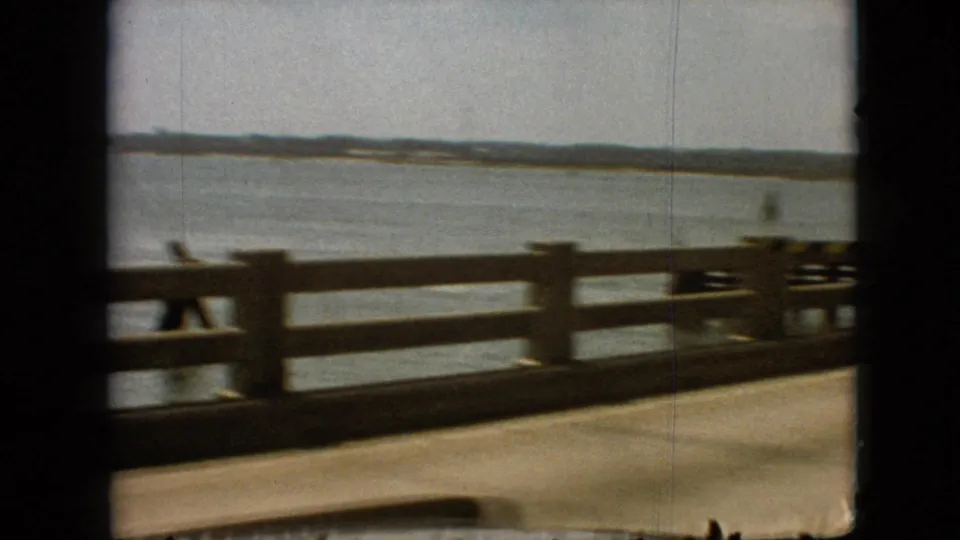 Moving car view across a lakeside pier or bridge, showing wooden railings, open water, and distant low hills in the 1968 Branson home-movie footage.