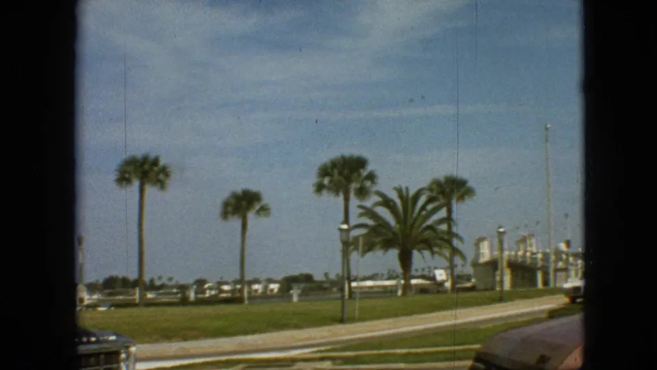 1968 Branson, Missouri 8mm frame showing palm trees, trimmed grass, low roadside structures, and the partial front of a classic car near the bottom edge.