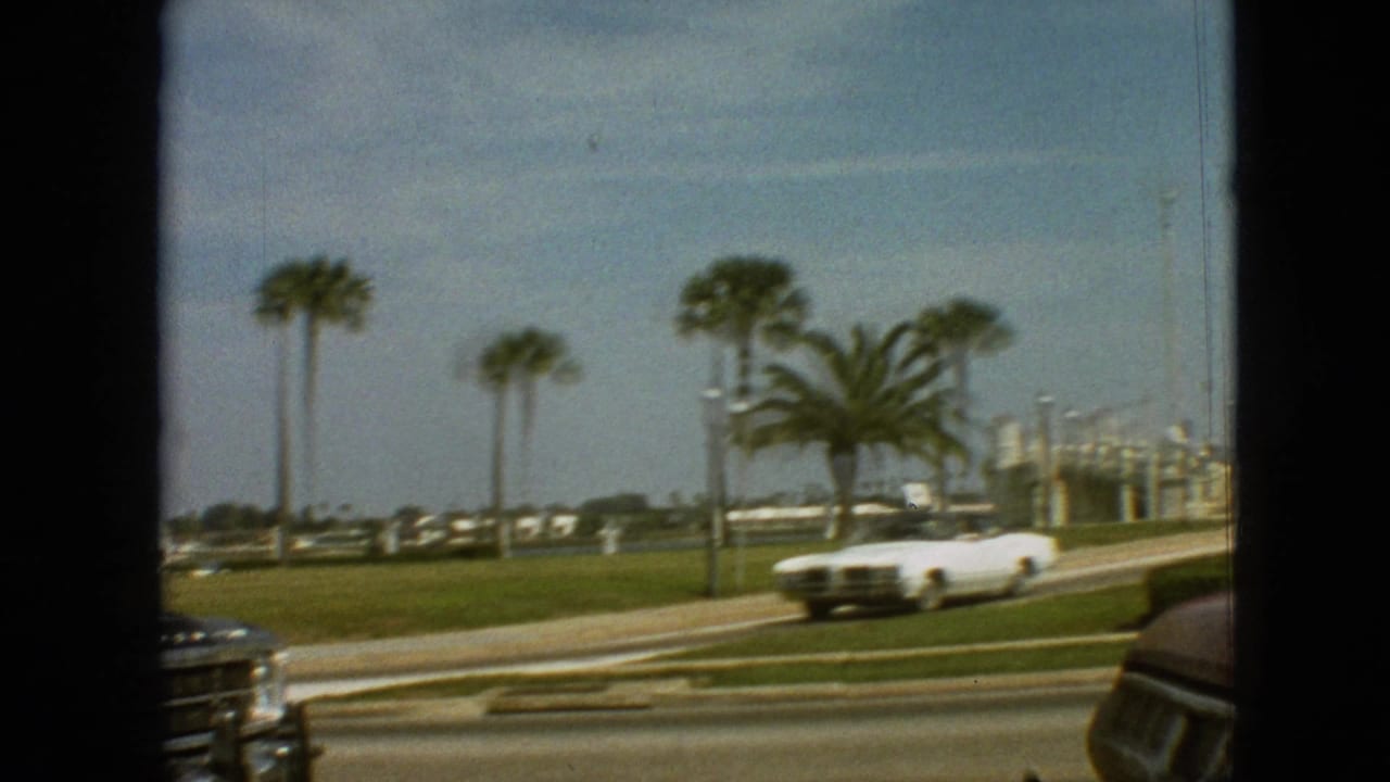 1968 Branson 8mm frame showing a white convertible on a palm-lined road, filmed from inside another moving vehicle.