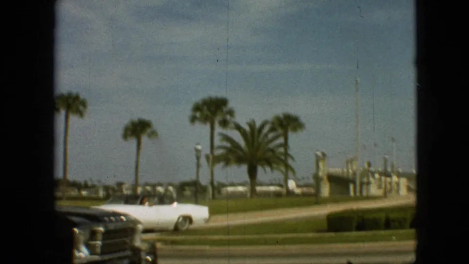 White convertible seen on a sunny Branson road in 1968, with palm trees, grass, and distant roadside structures behind it.
