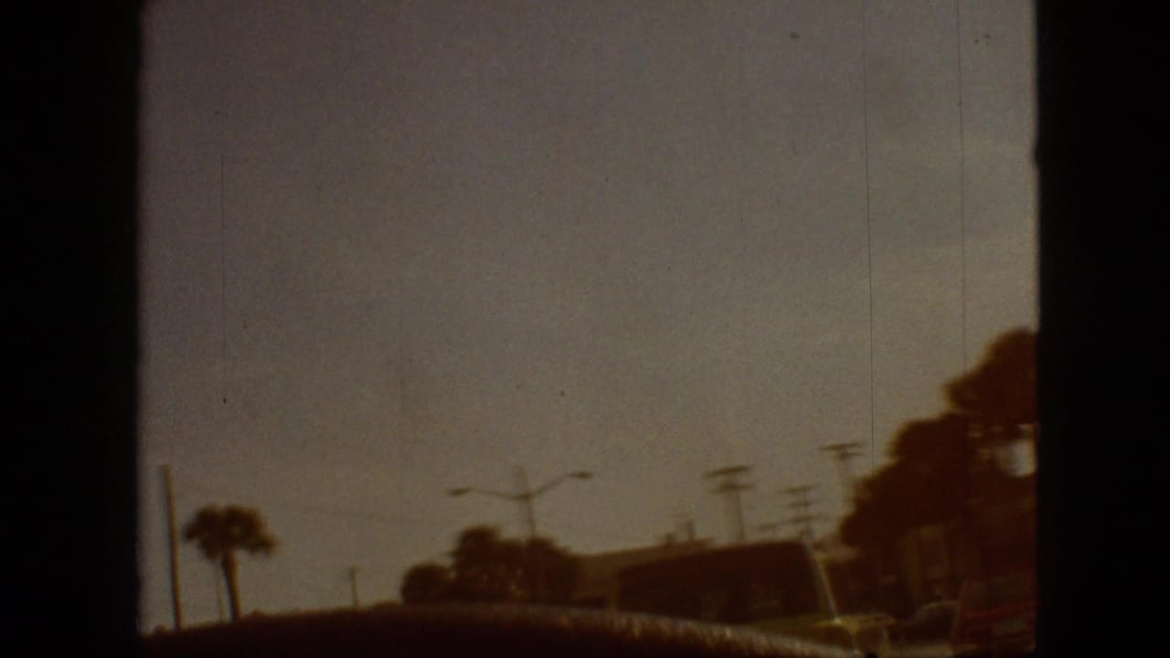 1968 home-movie frame with a dim passenger-side view past palm trees, utility poles, and roadside shapes, with heavy dark frame borders from the film scan.