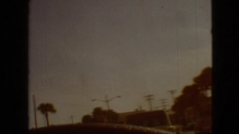 1968 home-movie frame with a dim passenger-side view past palm trees, utility poles, and roadside shapes, with heavy dark frame borders from the film scan.