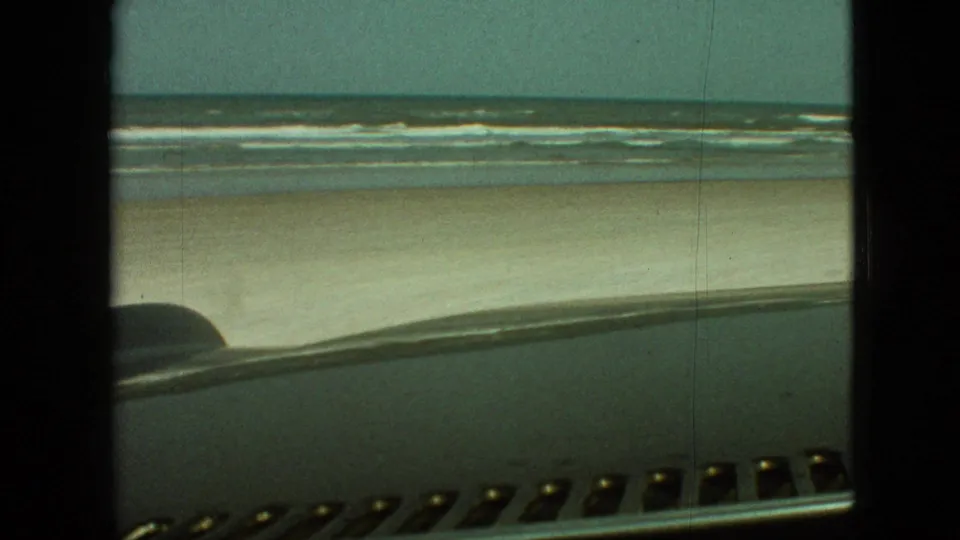 Wide 1968 home-movie view of an open sandy beach and rolling surf, framed from inside a car with the dashboard and windshield edge in the foreground.