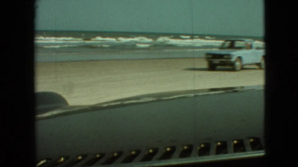 1968 home-movie frame of a light-colored vintage pickup moving across a sandy beach near the surf, seen from inside another car with the dashboard in front.