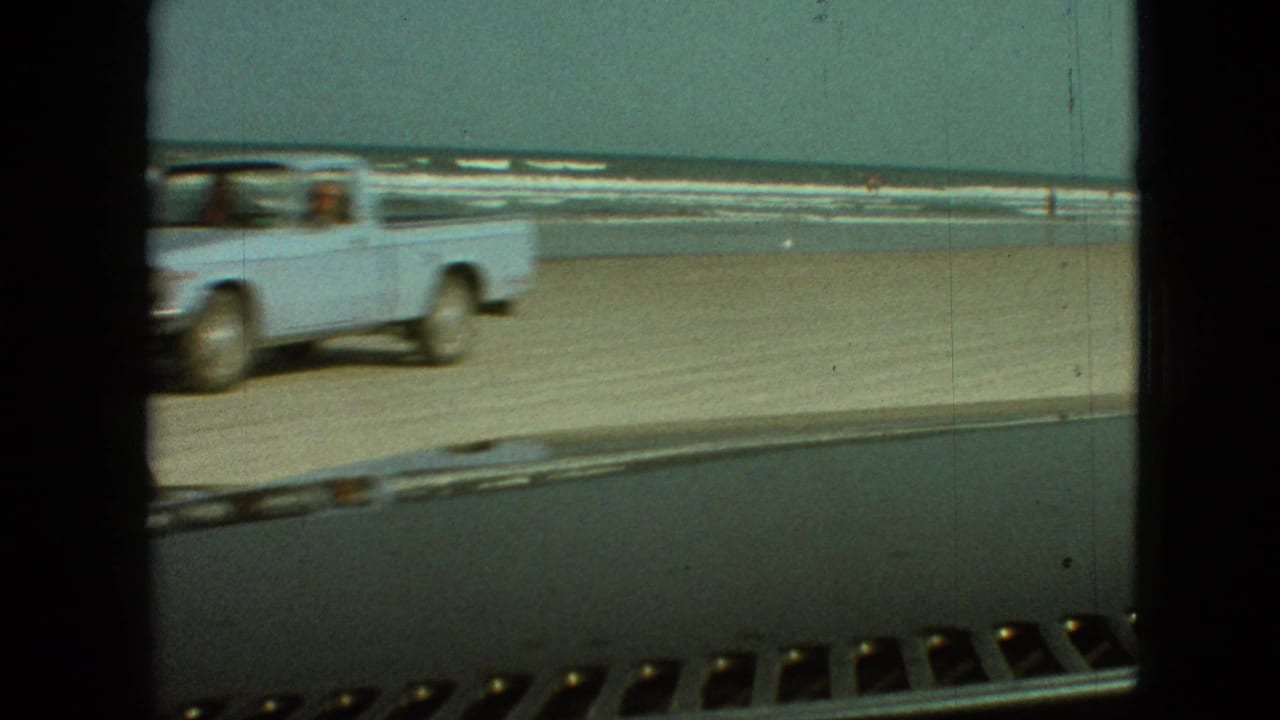 A light-colored pickup truck drives across a sandy beach near surf, seen from inside another vehicle in archival 8mm home-movie footage.