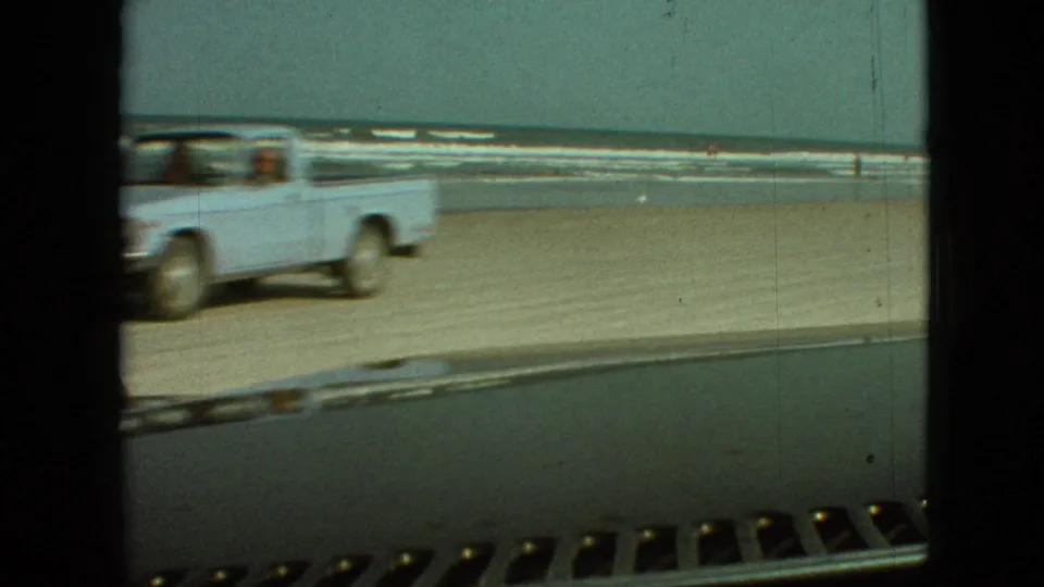 A light-colored pickup truck drives across a sandy beach near surf, seen from inside another vehicle in archival 8mm home-movie footage.