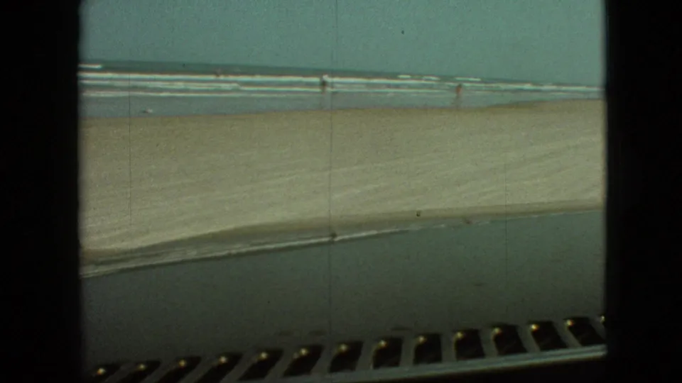 Open beach, ocean waves, and a distant figure are seen from a moving vehicle, with dashboard and film-frame edges visible in the scanned 8mm footage.