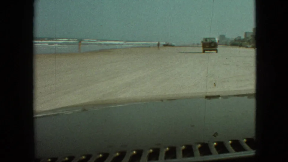 A pickup truck is seen farther ahead on the sandy shoreline, with surf to the left and small figures near the water in a 1960s-era home-movie frame.