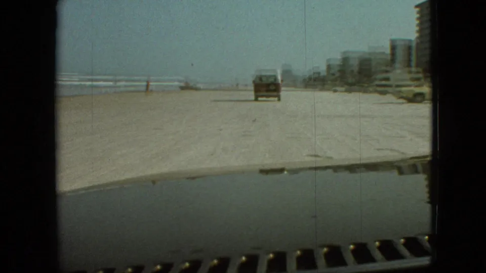 The vehicle follows a pickup truck along the beach, with surf, distant people, beachfront buildings, and parked cars visible in the background.