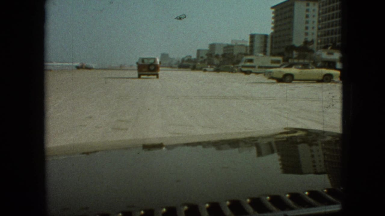 1968 8mm travel frame from the clip showing a sandy beach-like roadway, parked cars, a yellow convertible, mid-century shoreline buildings, and a vehicle hood reflection in the foreground.