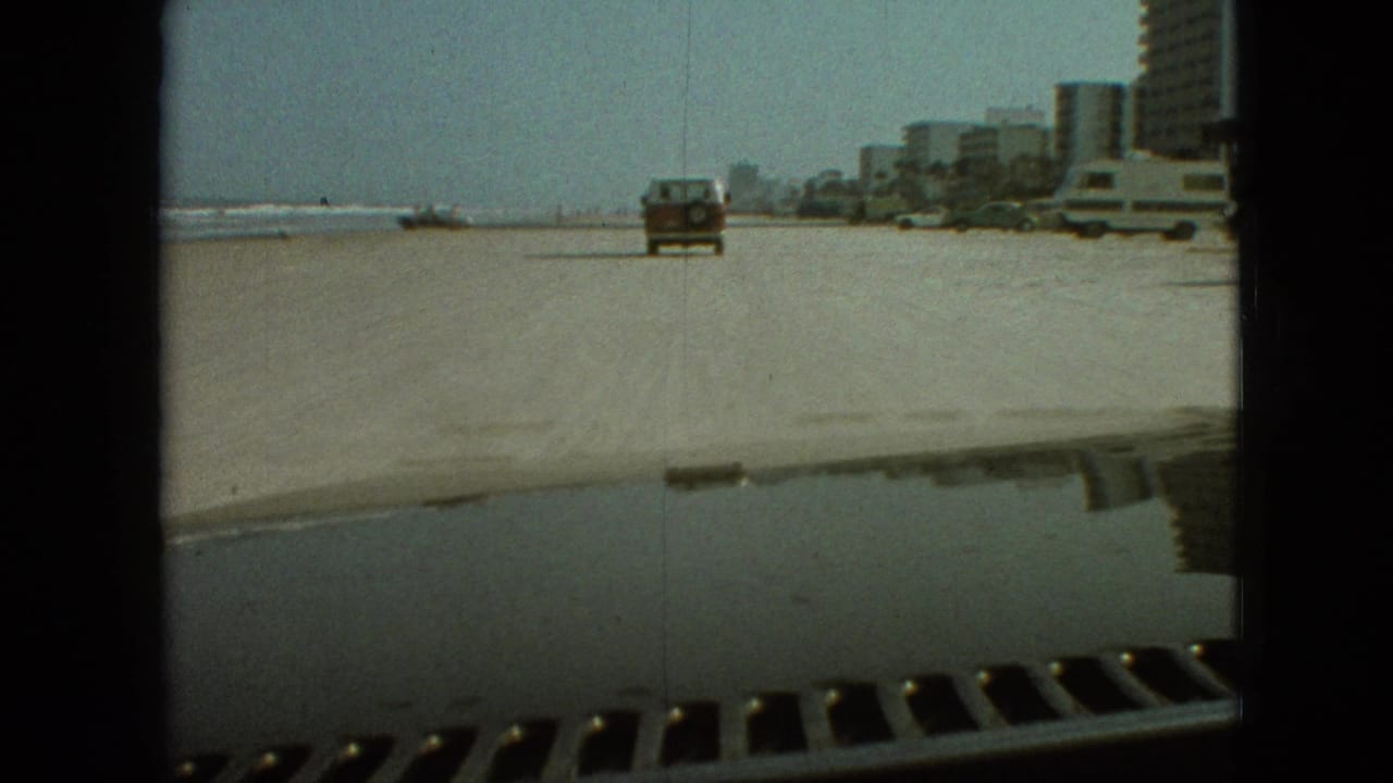 A red station wagon moves away from the camera on a wide sandy road, with shoreline buildings and parked vehicles in the distance during the 1968 home-movie travel sequence.
